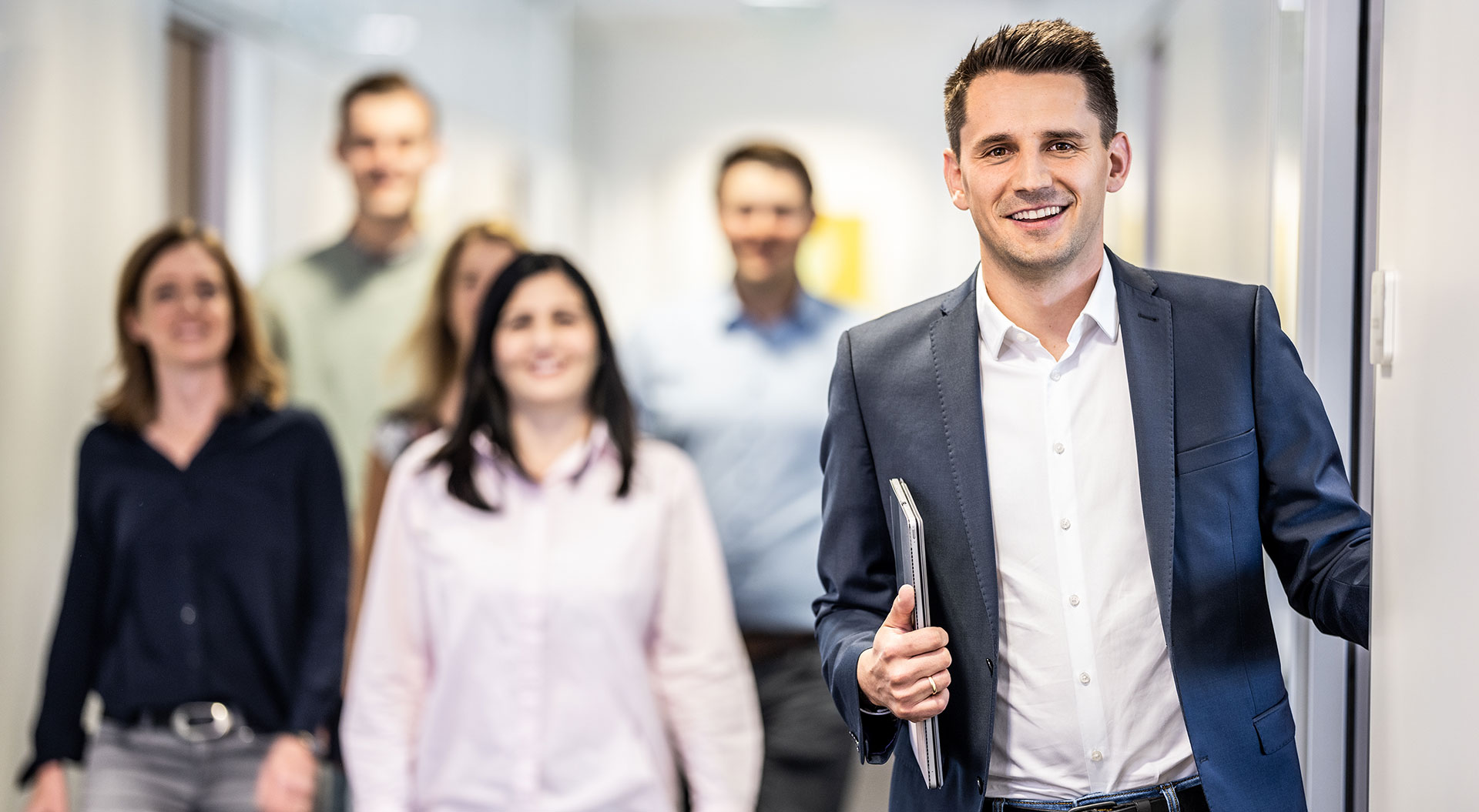 bjb-innovationen-mann-im-anzug-mit-laptop-in-der-hand-team-dahinter